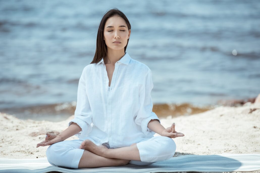 Theta Reprogramming focused woman meditating in ardha padmasana (half lotus pose) on yoga mat by sea